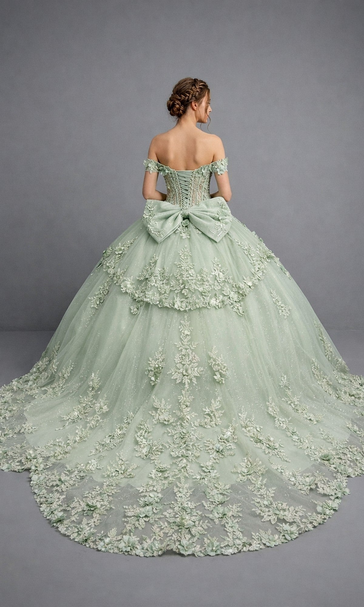 A woman stands facing away, wearing the Quinceanera Dress 5184 by Dancing Queen—a pastel green off-the-shoulder gown featuring intricate floral appliques, a lace-up back, and a voluminous ballgown skirt adorned with a large bow—set against a plain gray backdrop.